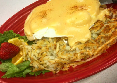 a plate with hash browns, two english muffins topped with fried eggs and hollandaise sauce with fruit garnish on the side