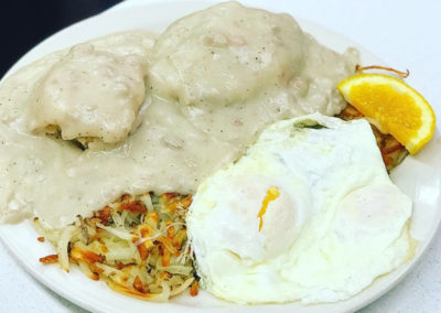 a plate with hash browns, two biscuits smothered in sausage gravy, fried eggs and an orange slice for garnish